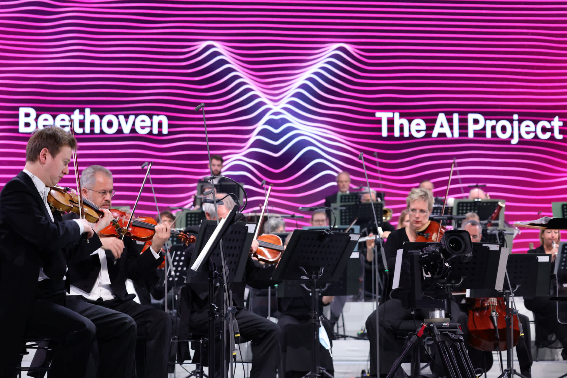 An orchestra playing in front of a large digital display that says, â€œBeethoven X  The AI Projectâ€�