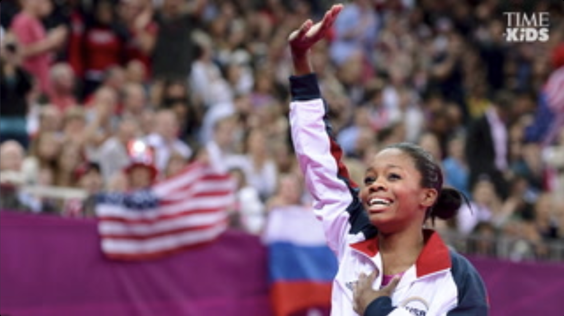 Gabby Douglas smiling, with one hand up above her head and one hand on her chest, against a backdrop of a stadium with an American flag.