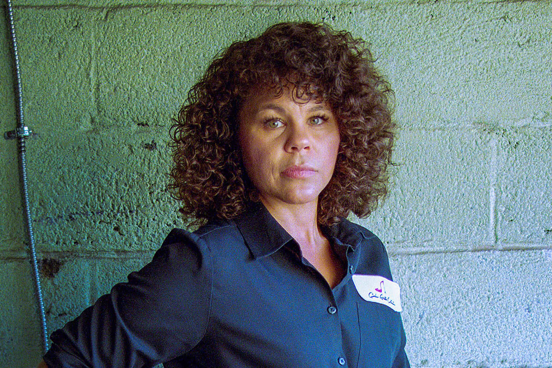 A woman wearing a dark blue shirt stands against a cinderblock wall.