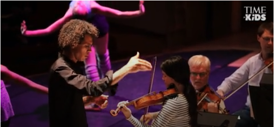 A conductor and violinists practicing with a dancer practicing in the background