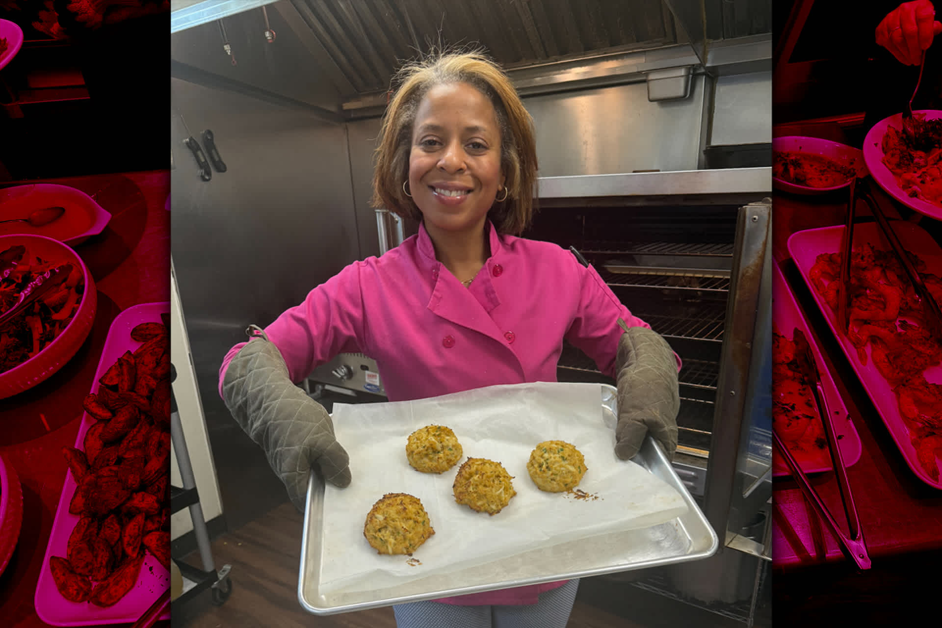 A woman wearing a pink chef jacket and gray oven mitts takes a tray of baked goods fresh from the oven.