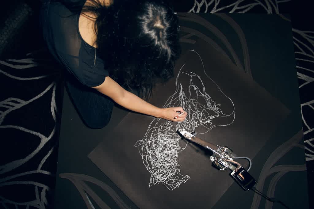 An artist and a robotic arm draw with white pens on a black sheet of paper.