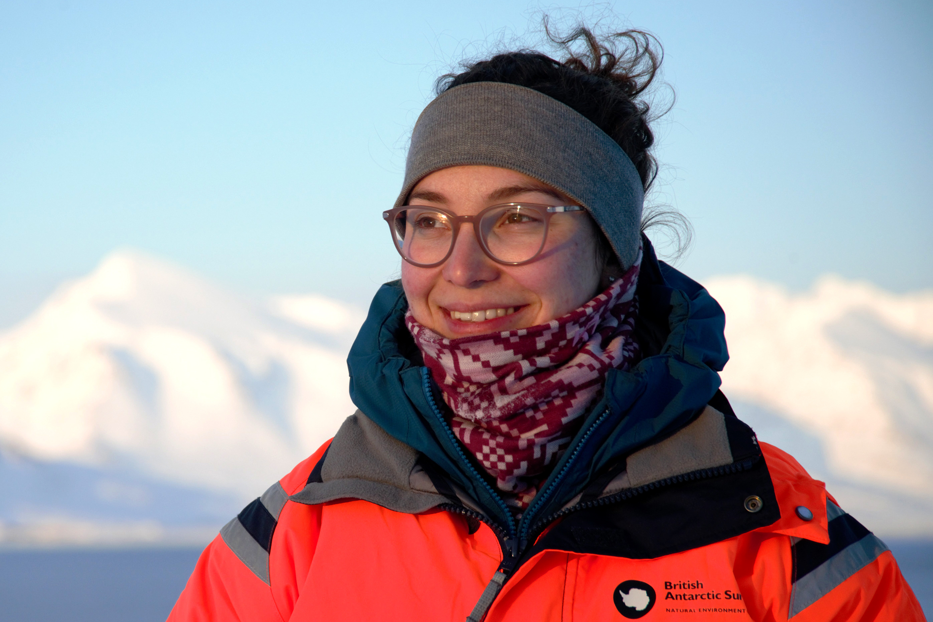 A scientist wearing a bright orange jacket with a British Antarctic Survey patch stands outdoors in a snowy, icy landscape. 