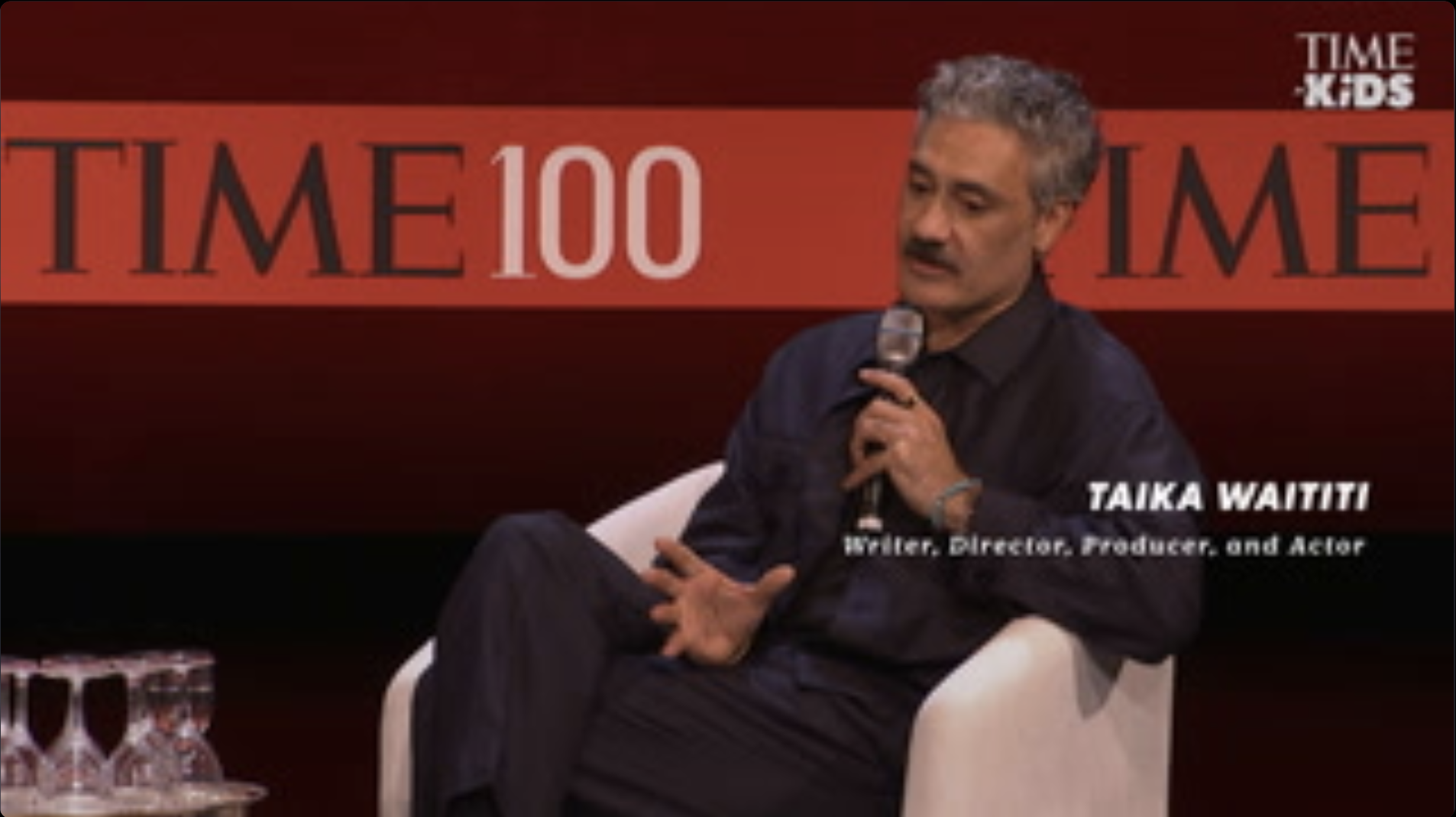 Taika Waititi, seated in a chair with a microphone to his mouth. 