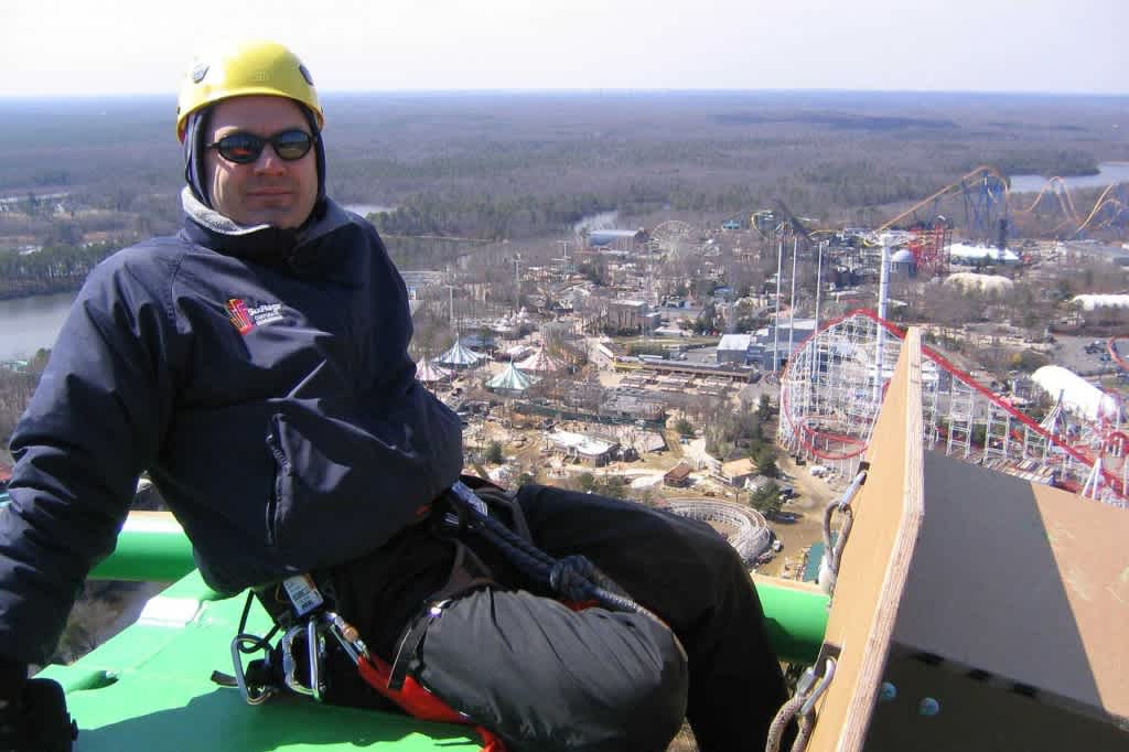Larry Chickola sits at the peak of a rollercoaster track wearing a helmet, sunglasses and climbing harness