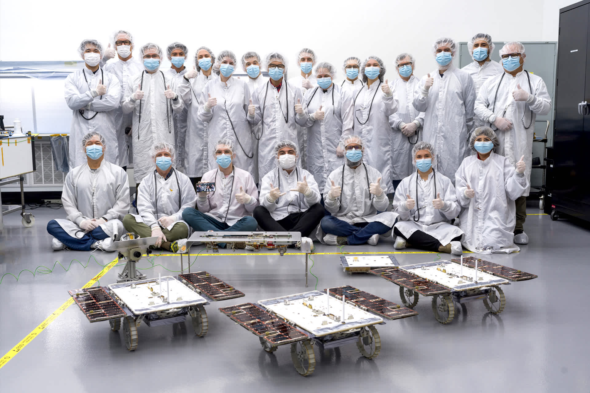 A group poses with thumbs-up behind three rovers.