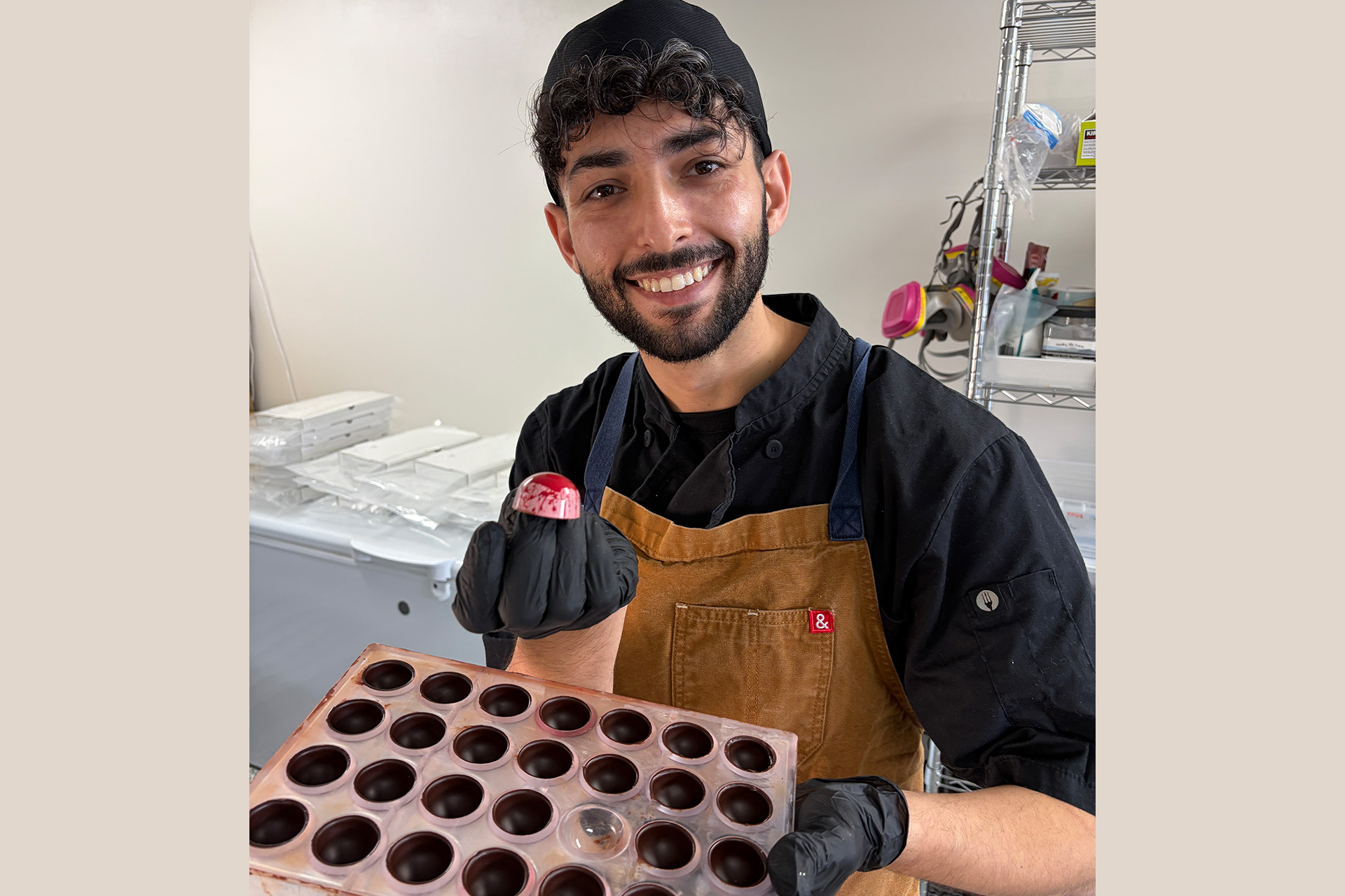 A chocolatier smiles while holding a tray of chocolate shells and a finished pink chocolate candy.