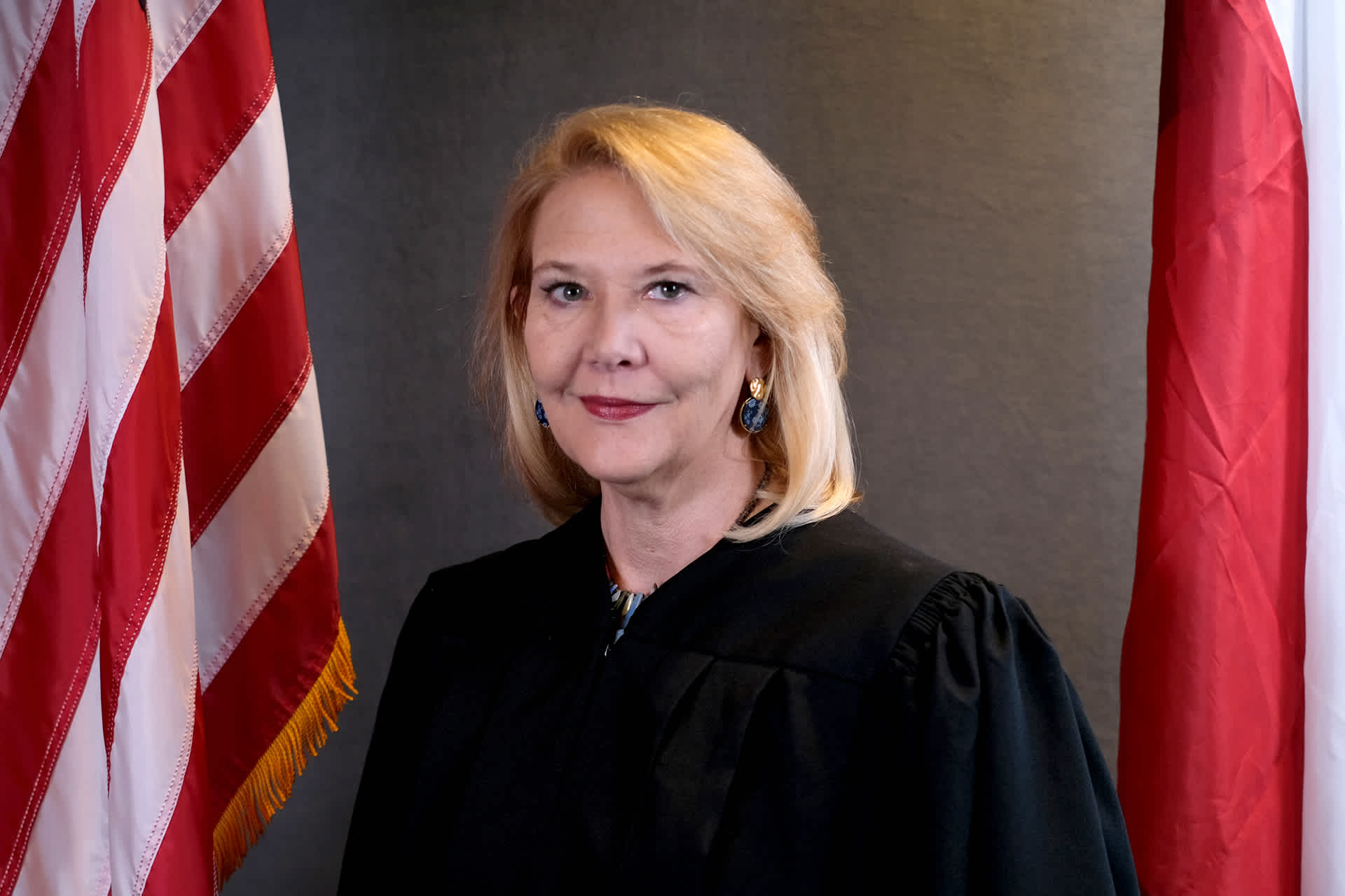 A female judge with blonde hair poses between two flags.