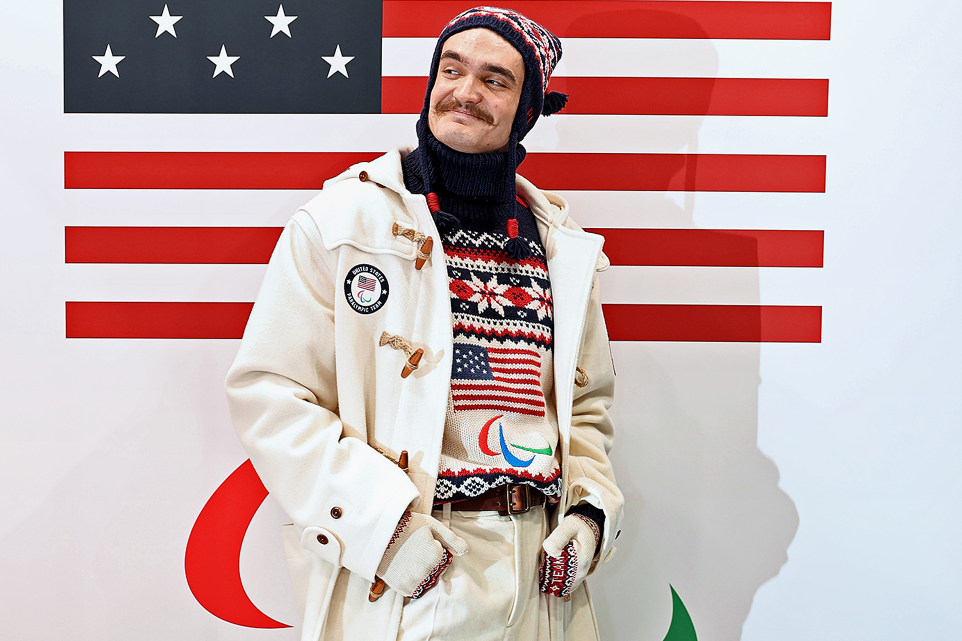 A man wearing winter clothing and a knit hat stands in front of a large flag.