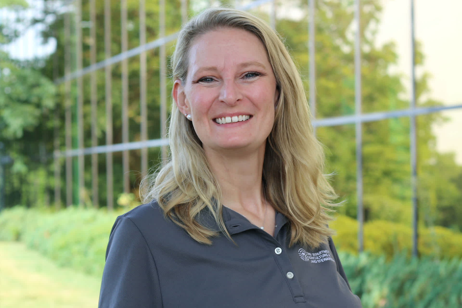 A woman in a gray polo-style shirt smiles as she poses outdoors.
