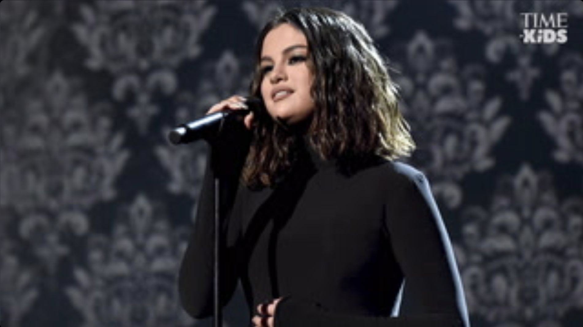 Selena Gomez standing in front of a mic stand with one hand on the mic and the other placed on her stomach.