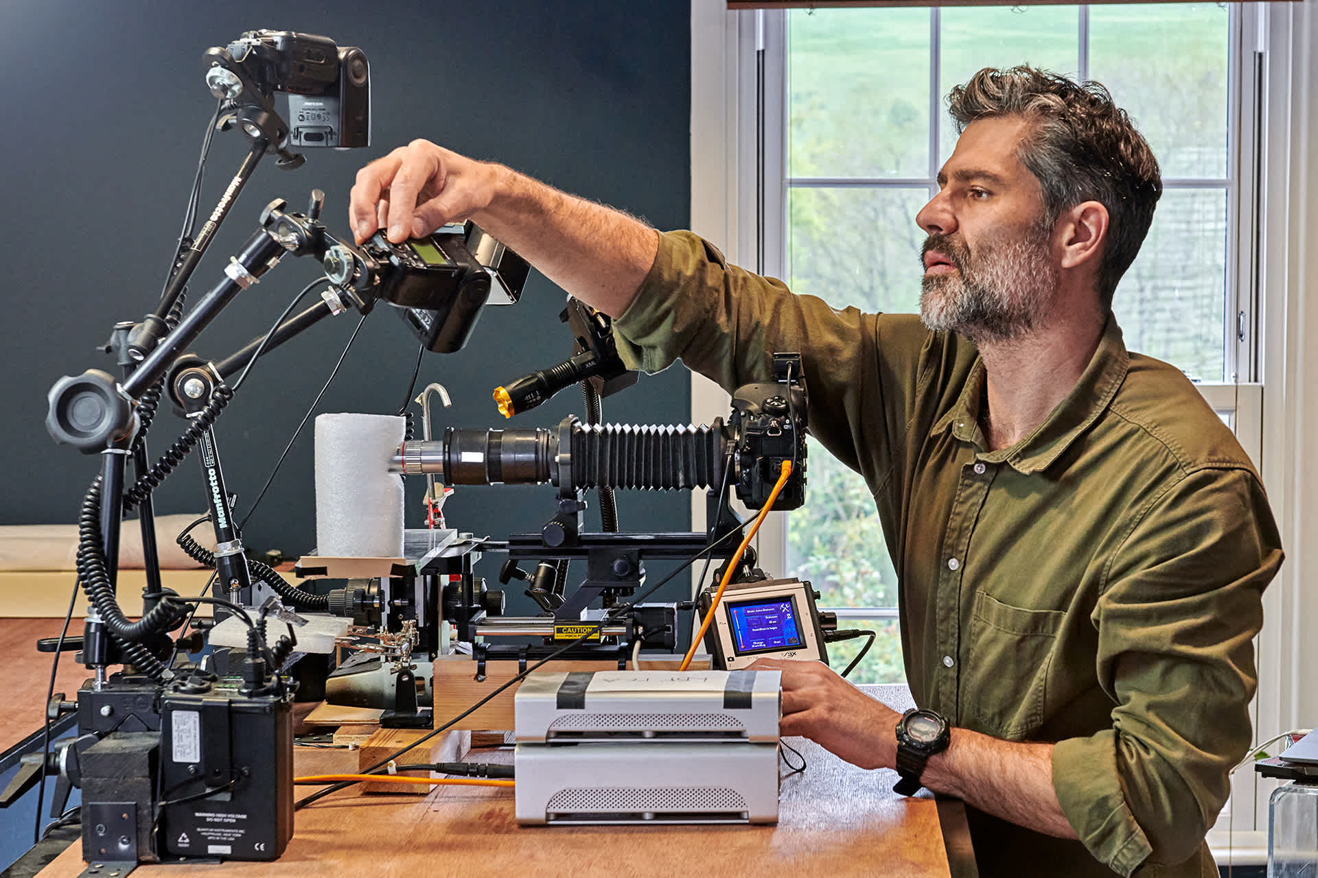 A man sets up a camera in front of a window in a blue room.