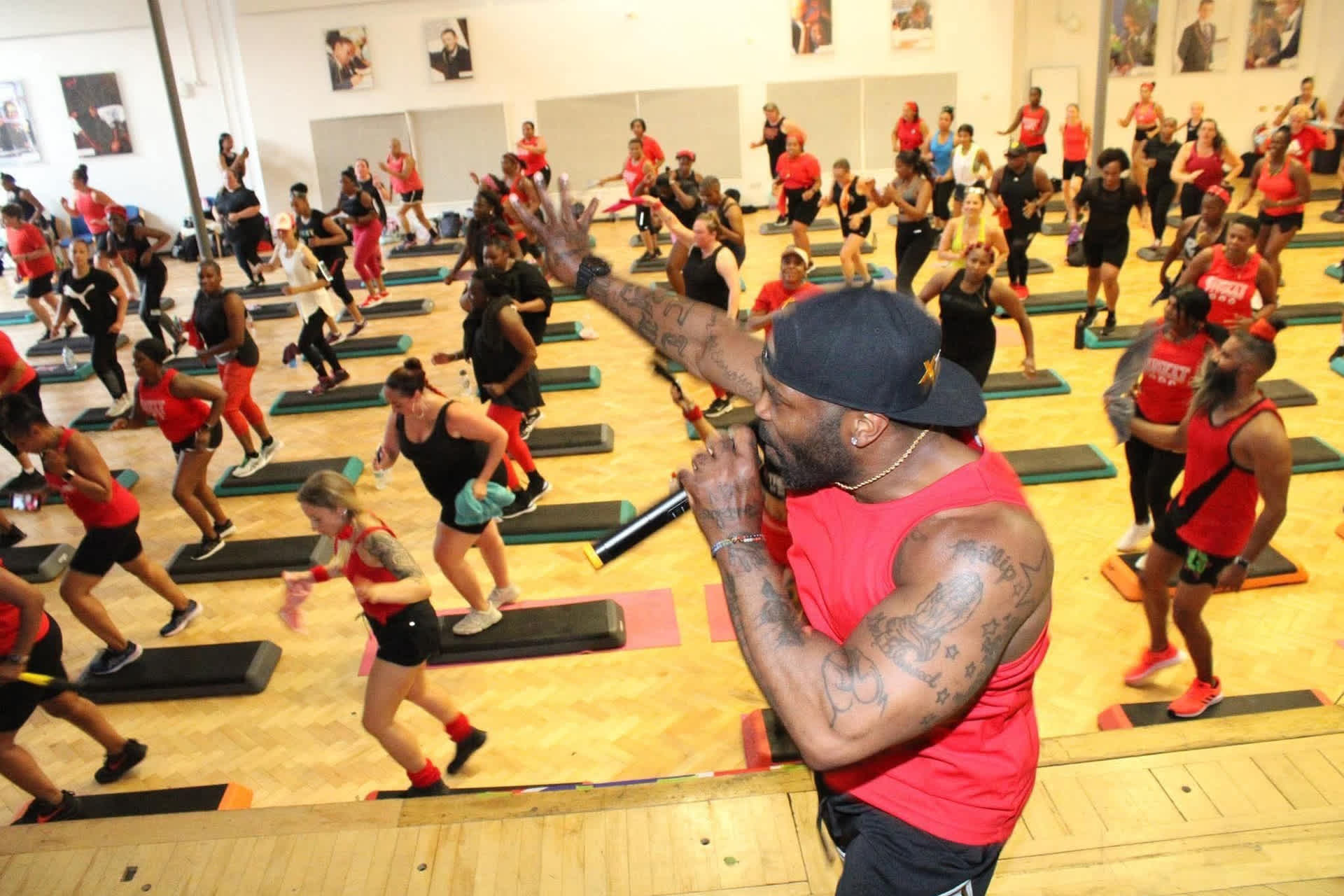 A man with a red sleeveless top and black baseball cap speaks into a microphone as he leads a workout for a large group of people.