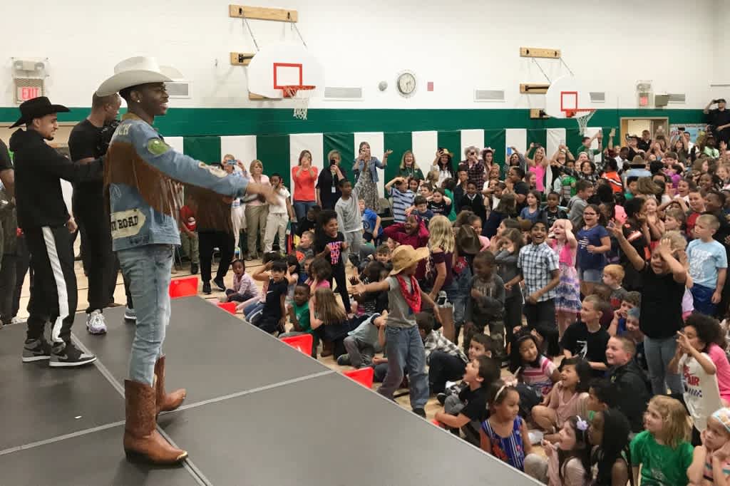 Musician Lil Nas X performs in a school gymnasium in front of a group of children.