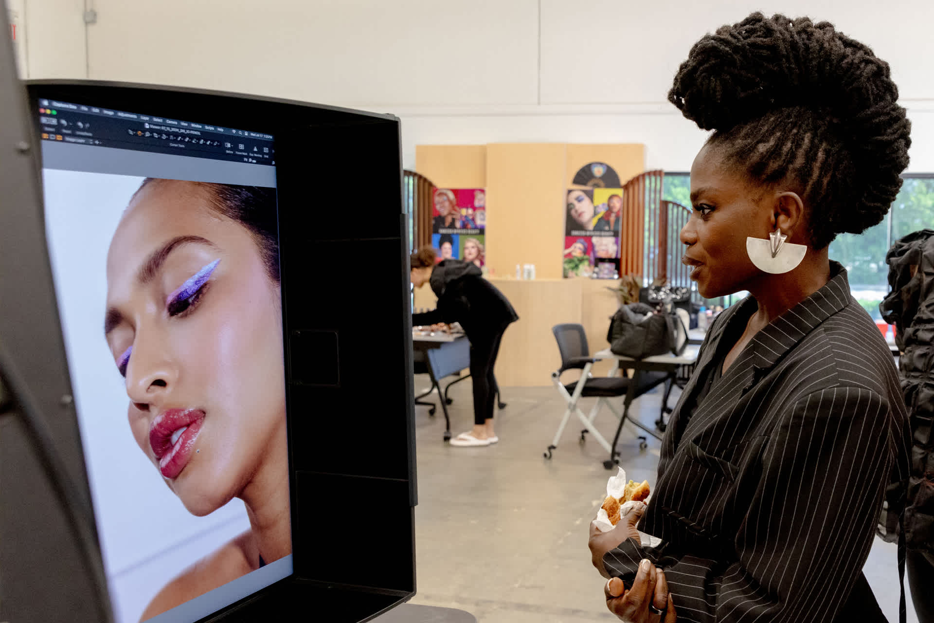 A woman stands in front of a monitor, looking at a photo of a woman wearing colorful makeup.