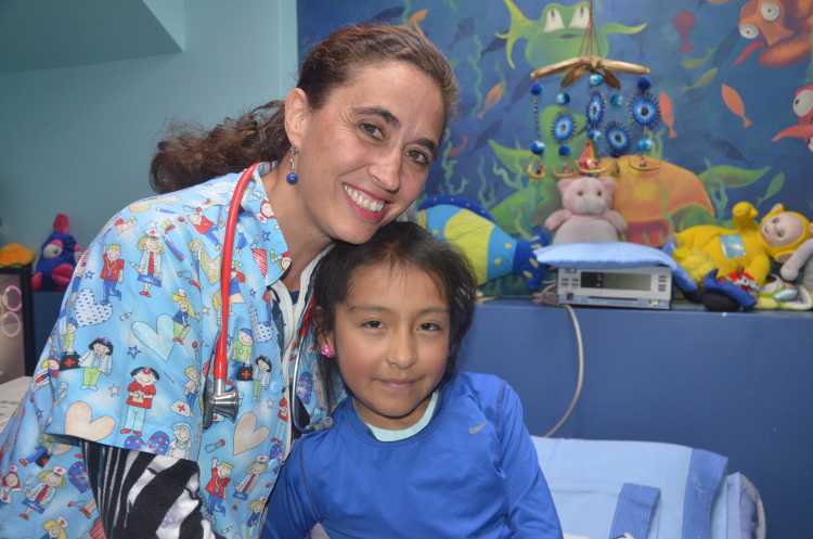 A doctor in colorful scrubs smiles with a young patient on a hospital bed