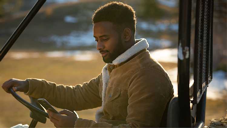 A man wearing a tan shearling jacket sits in a utility vehicle outdoors, looking at his smartphone with a slight smile, during a sunny winter day.