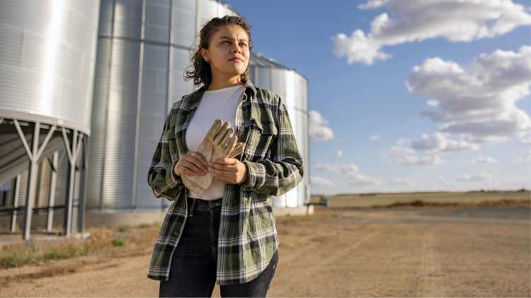 Une personne portant une chemise verte à carreaux tient des gants de travail devant des silos à grains.