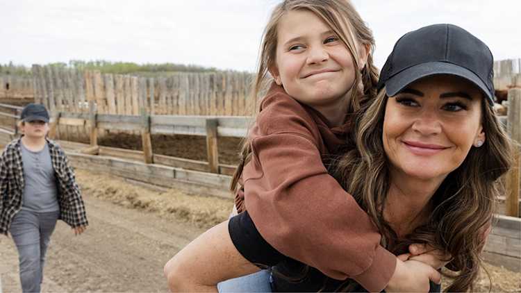 Femme marchant dans une cour de ferme avec un enfant sur son dos. Tous deux sourient.