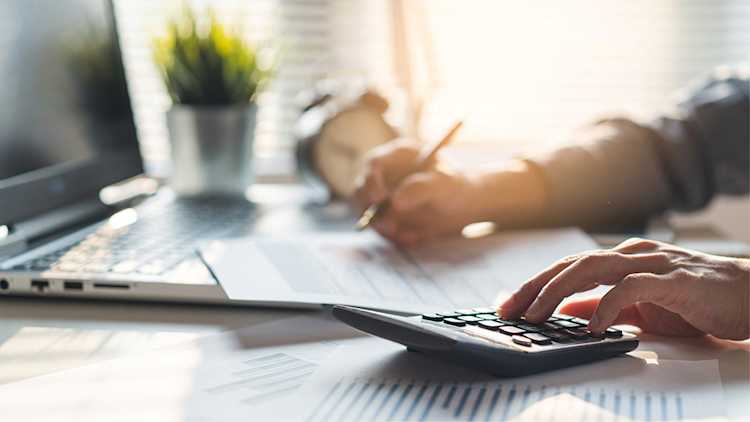 Person working at a laptop with a calculator and sheet of paper.