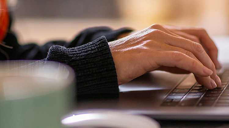 Close-up of a person's hands typing on a laptop keyboard in a softly lit setting, with a blurred mug in the foreground.