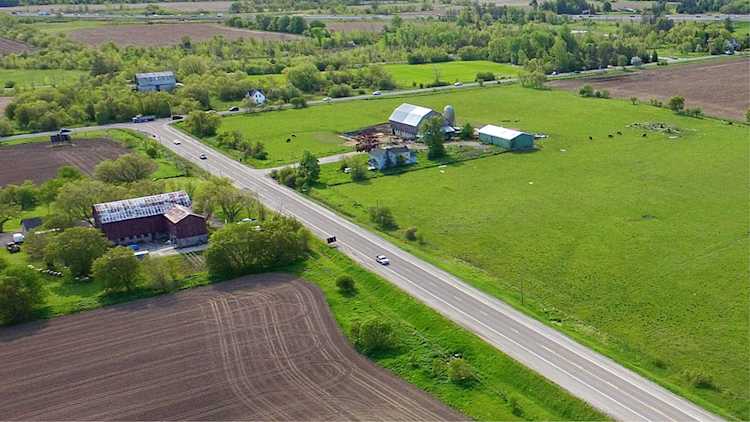 Aerial view of a rural farming landscape with barns, fields, scattered houses, and a paved road running through green pastures and cultivated land on a sunny day.