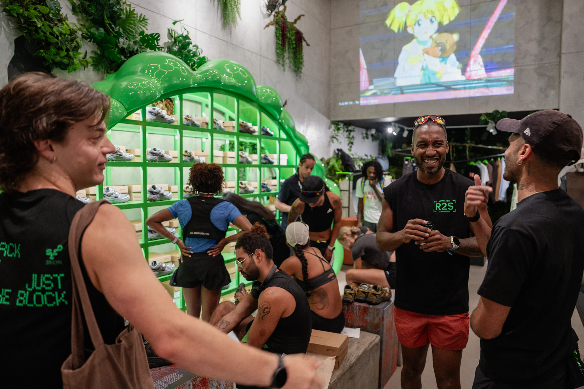 People gathered in a modern retail store with green shelving displaying sneakers, plants, and a large screen above.