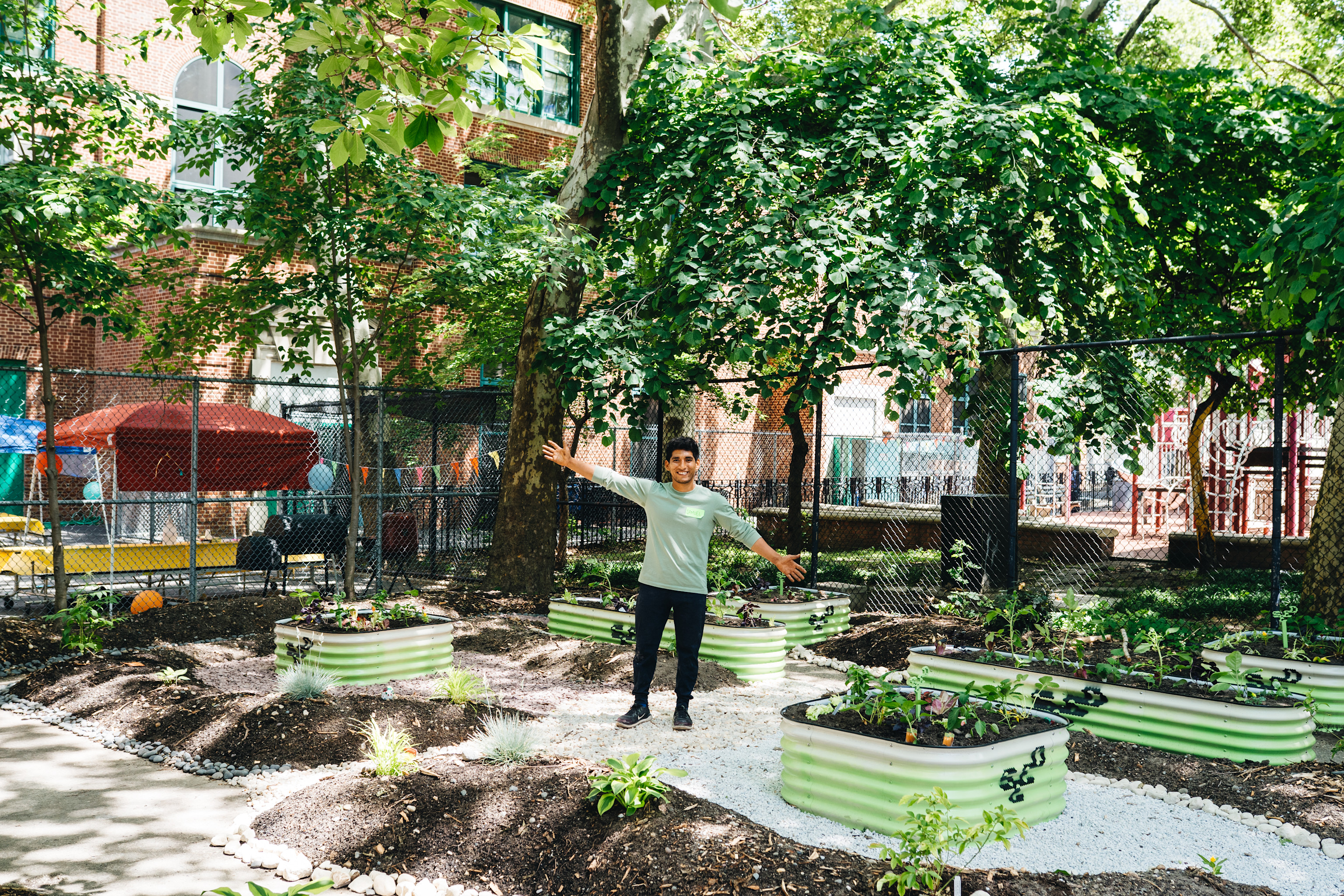 Person standing in community garden with raised planter beds, surrounded by trees and urban buildings in background.