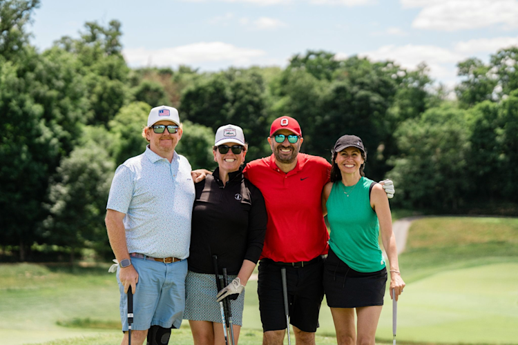 Four golfers in casual attire and caps pose together on a golf course with lush green trees in the background.