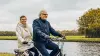 An elderly couple riding on a tandem bicycle along a river