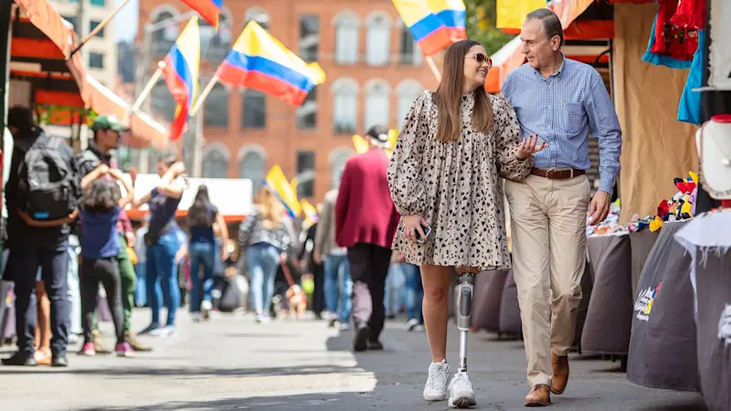 Una pareja caminando en terreno plano a través de un mercado al aire libre. Uno de ellos lleva la rodilla mecánica Ottobock Dynion