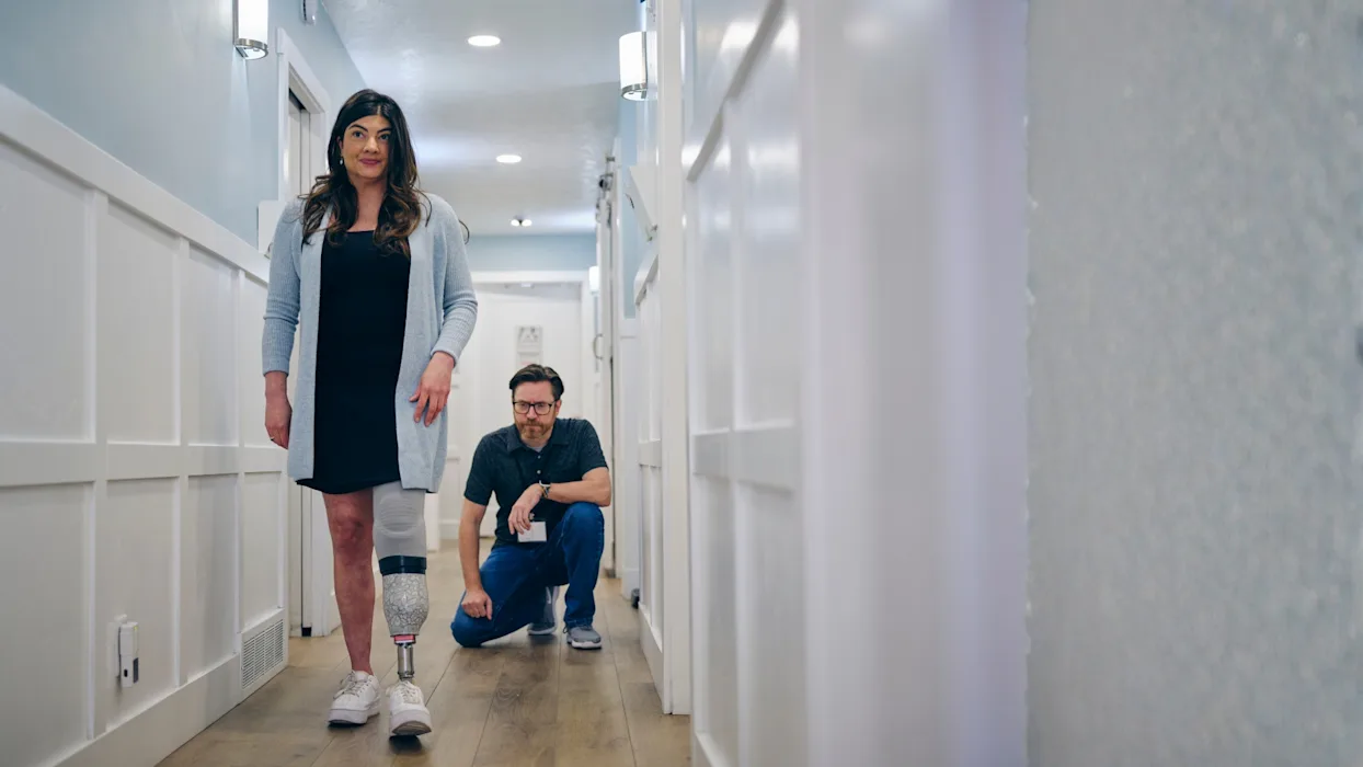 A lower limb prosthetic leg user walking in a hallway while their Certified Prosthetist Orthotist monitors their gait