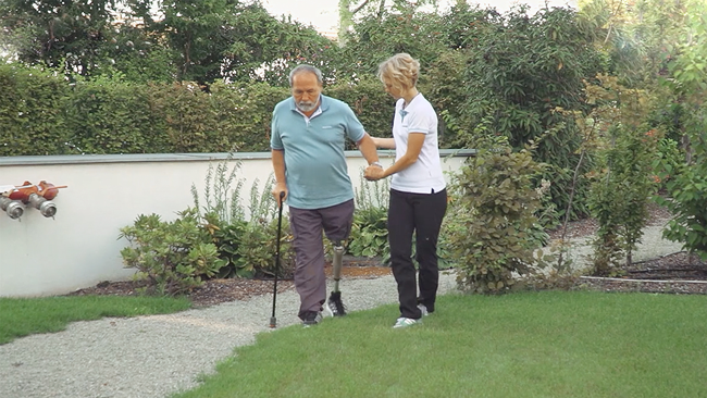 An elderly prosthetic leg user walking on an outdoor trail, accompanied by a Certified Prosthetist Orthotist
