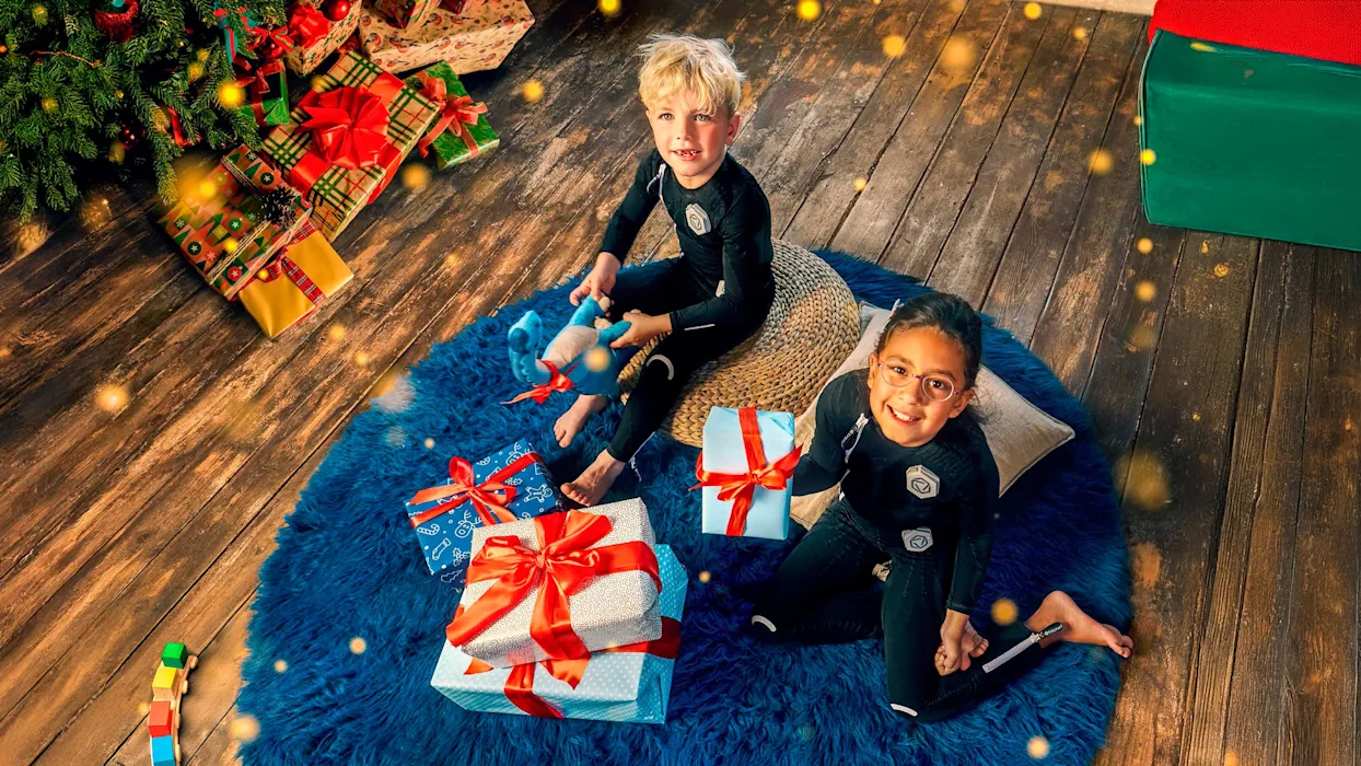 Due bambini che indossano la tuta Exopulse Mollii giocano con i pacchi dono sotto l'albero di natale
