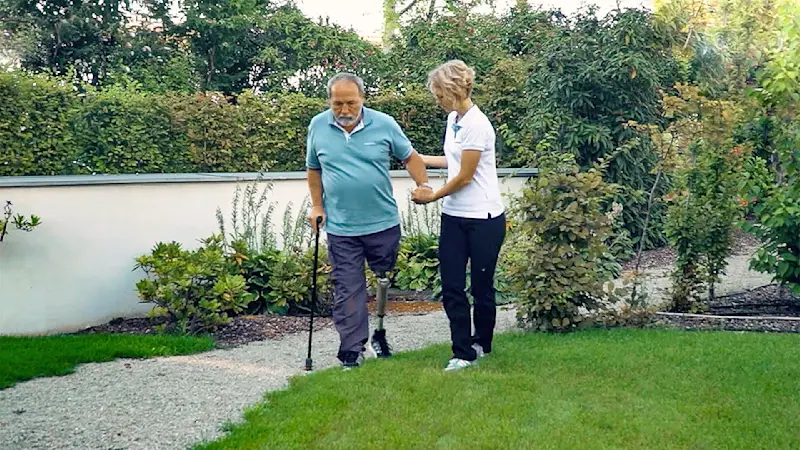 Un usuario de prótesis de rodilla Ottobock Kenevo caminando al aire libre en una pasarela de asfalto con asistencia