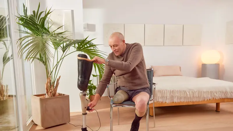 A lower limb amputee setting their Genium prosthetic leg aside as they sit in a chair in a dining room setting