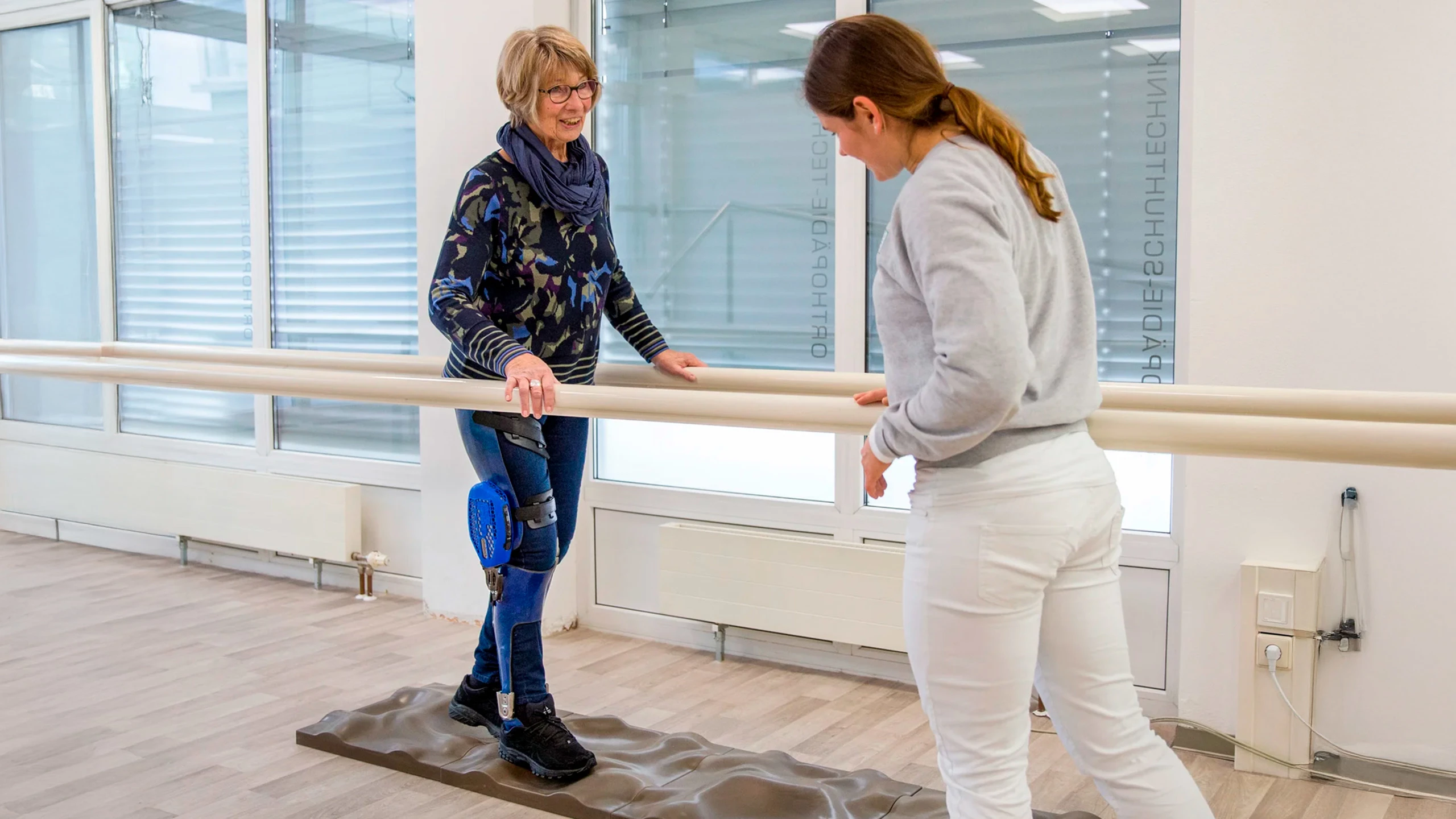 Un'utente anziana di C-Brace prova l'ortesi su una pedana che simula una superficie irregolare sotto lo sguardo attento della fisioterapista Ottobock.care