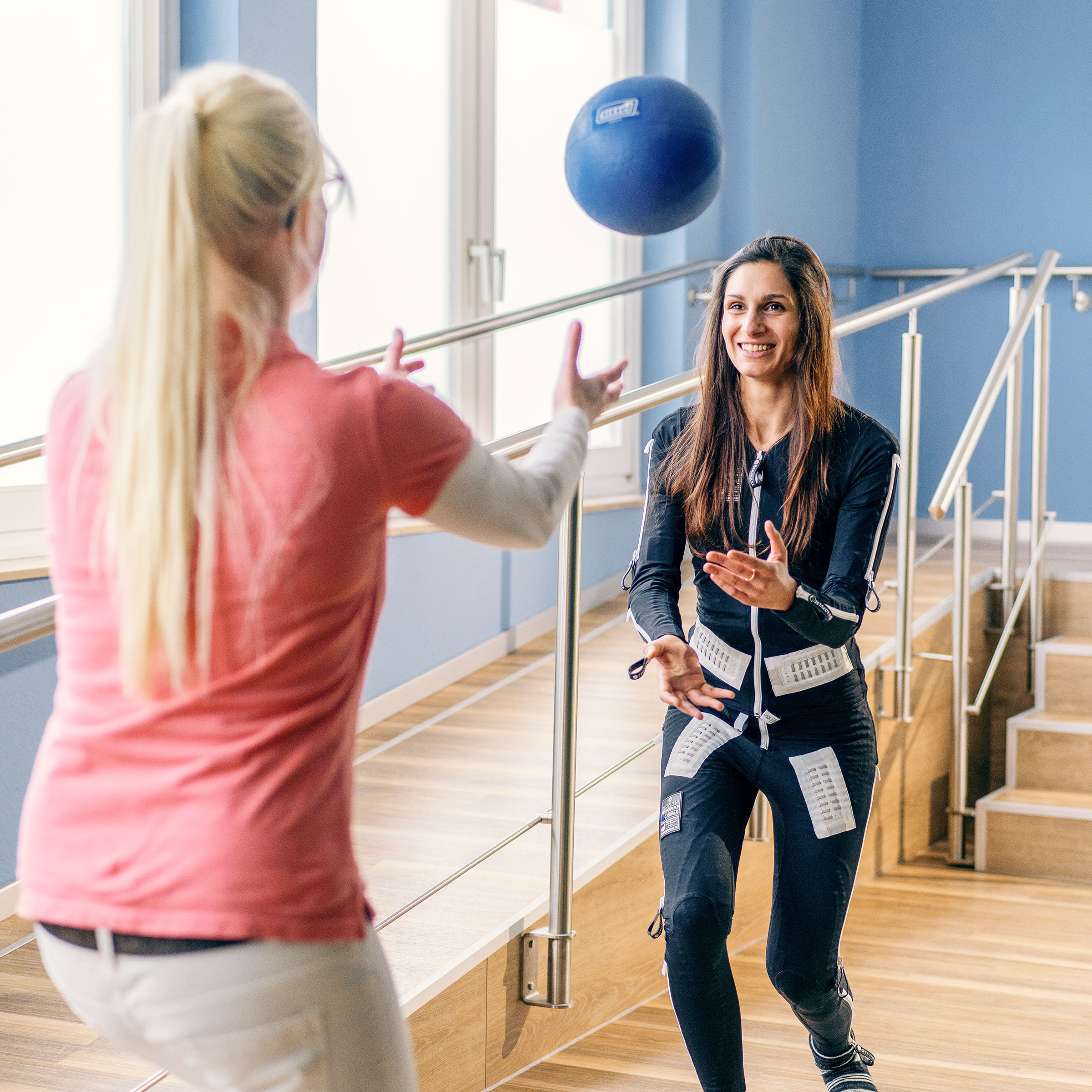 Louisa con la tuta Exopulse mentre fa esercizio in palestra con l'aiuto di una fisioterapista e una palla