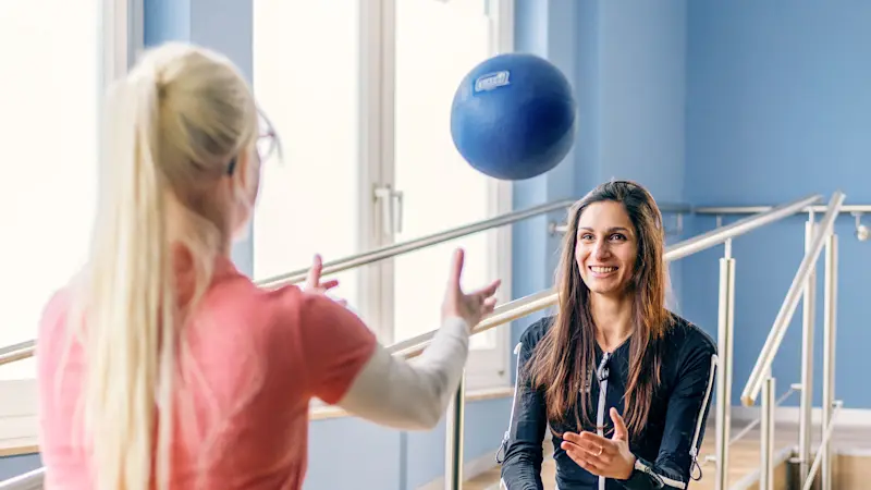 Louisa con la tuta Exopulse mentre fa esercizio in palestra con l'aiuto di una fisioterapista e una palla