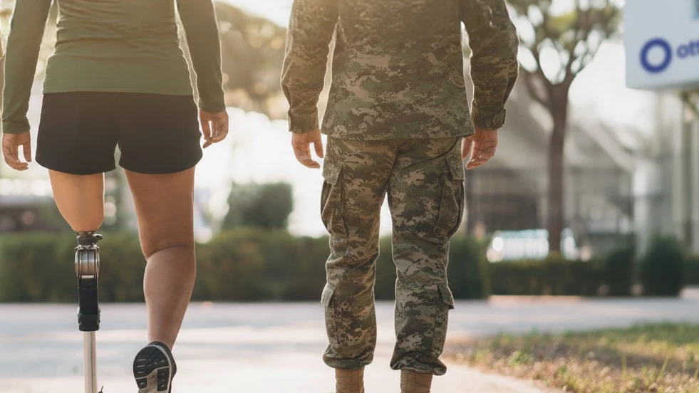 Two military veterans, one with limb difference, walking outdoors along a sidewalk