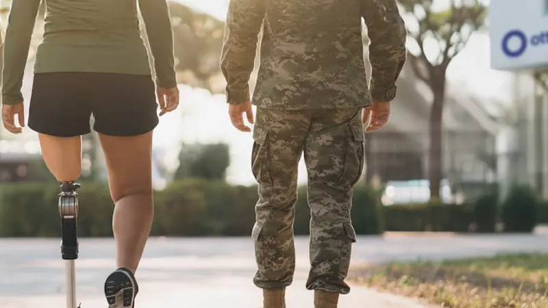 Two military veterans, one with limb difference, walking outdoors along a sidewalk