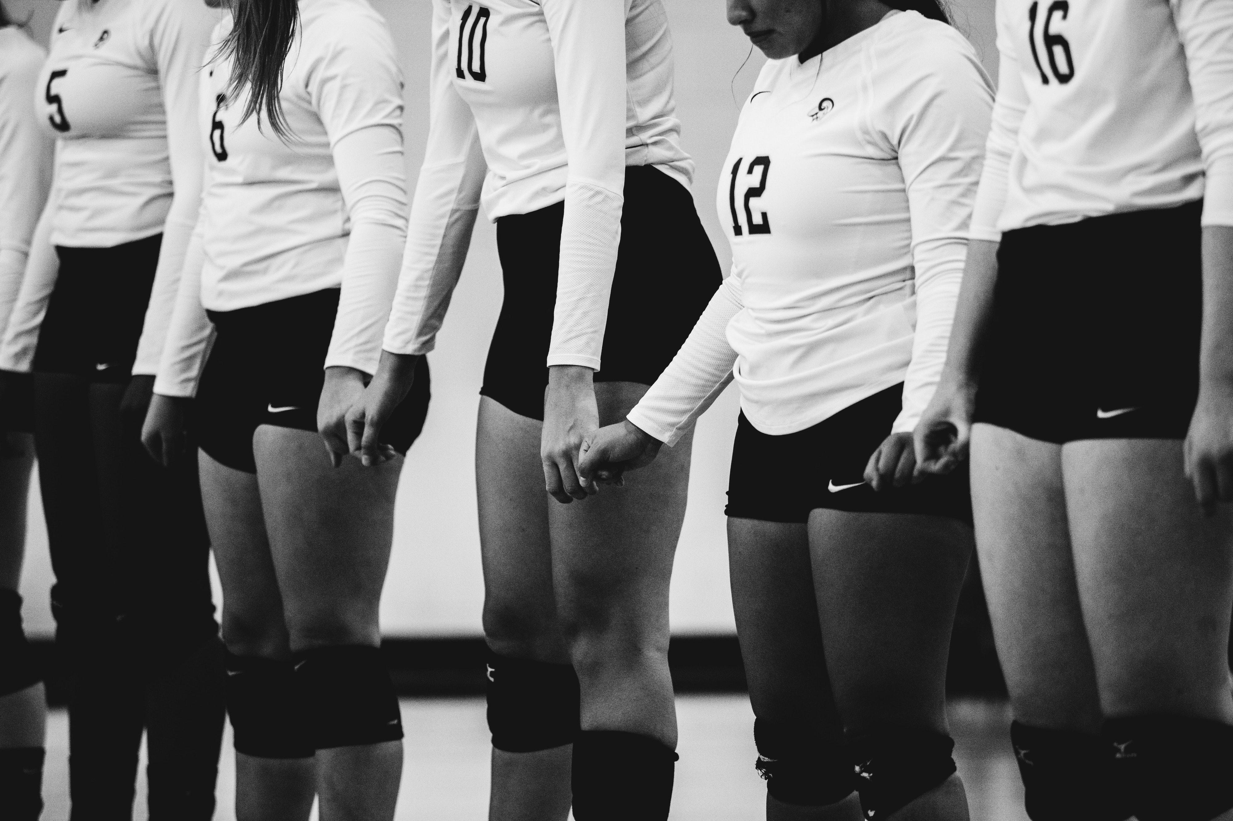 Five people, assumingly women, standing in line, holding hands. They are wearing sports clothes: Long-sleeved white shirts with numbers on the front of the shirts, and short black shorts with a Nike logo on them.