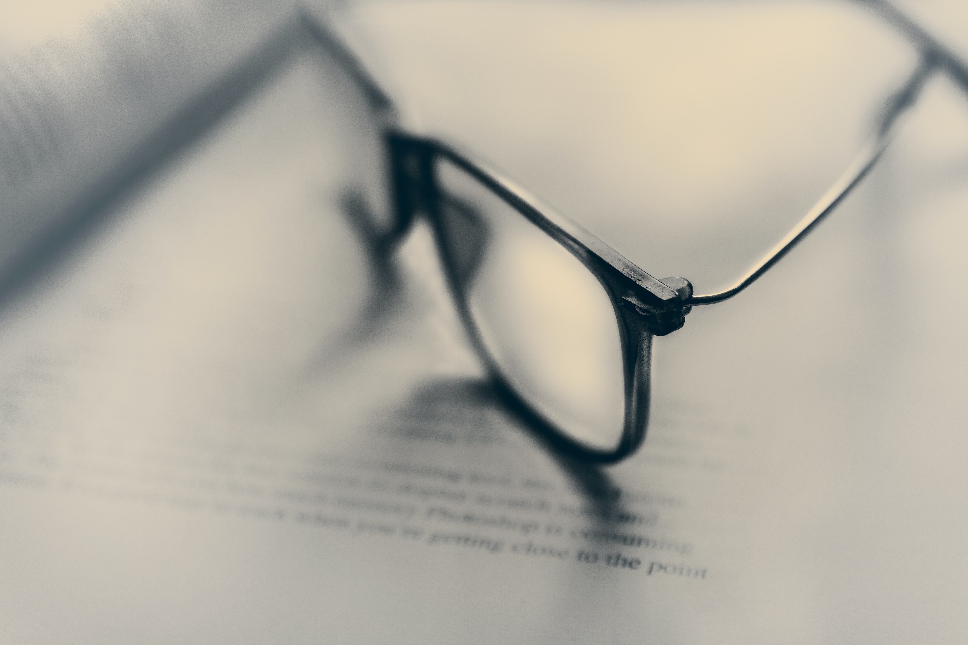 A blurred view with eyeglasses on a book. Text is unreadable, and the picture is grey-scaled.