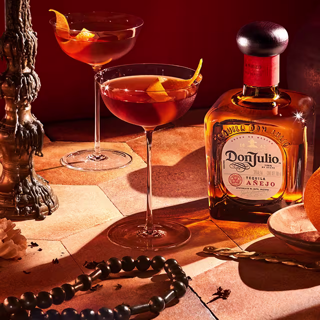 Two people holding glasses full of Añejo Manhattan on a table next to a bottle of Don Julio Añejo