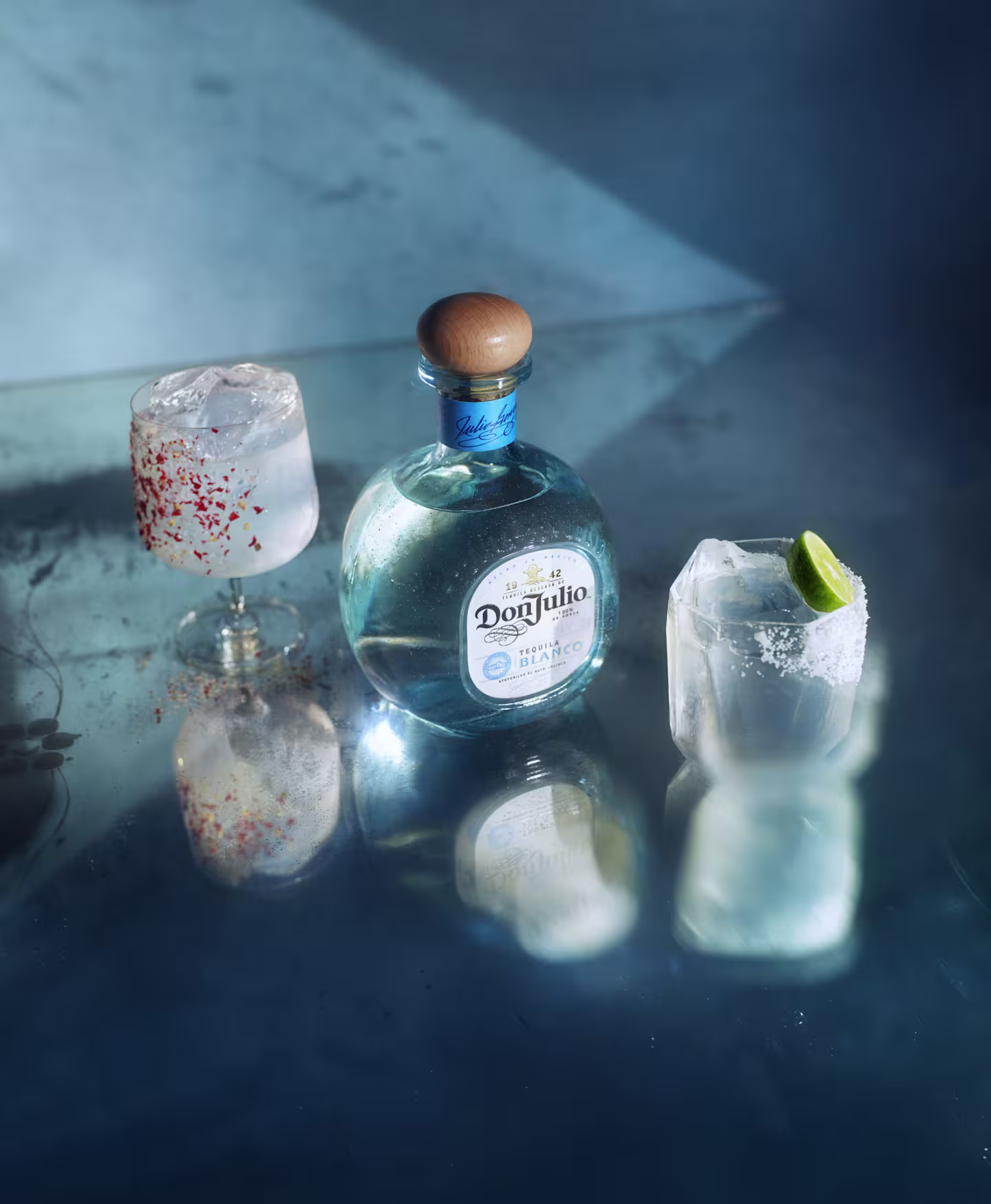 Image of a Don Julio Blanco tequila bottle on a reflective surface, flanked by two glasses with ice. One glass has chili flakes; the other is rimmed with salt and topped with a lime wedge, set against a cool blue background.
