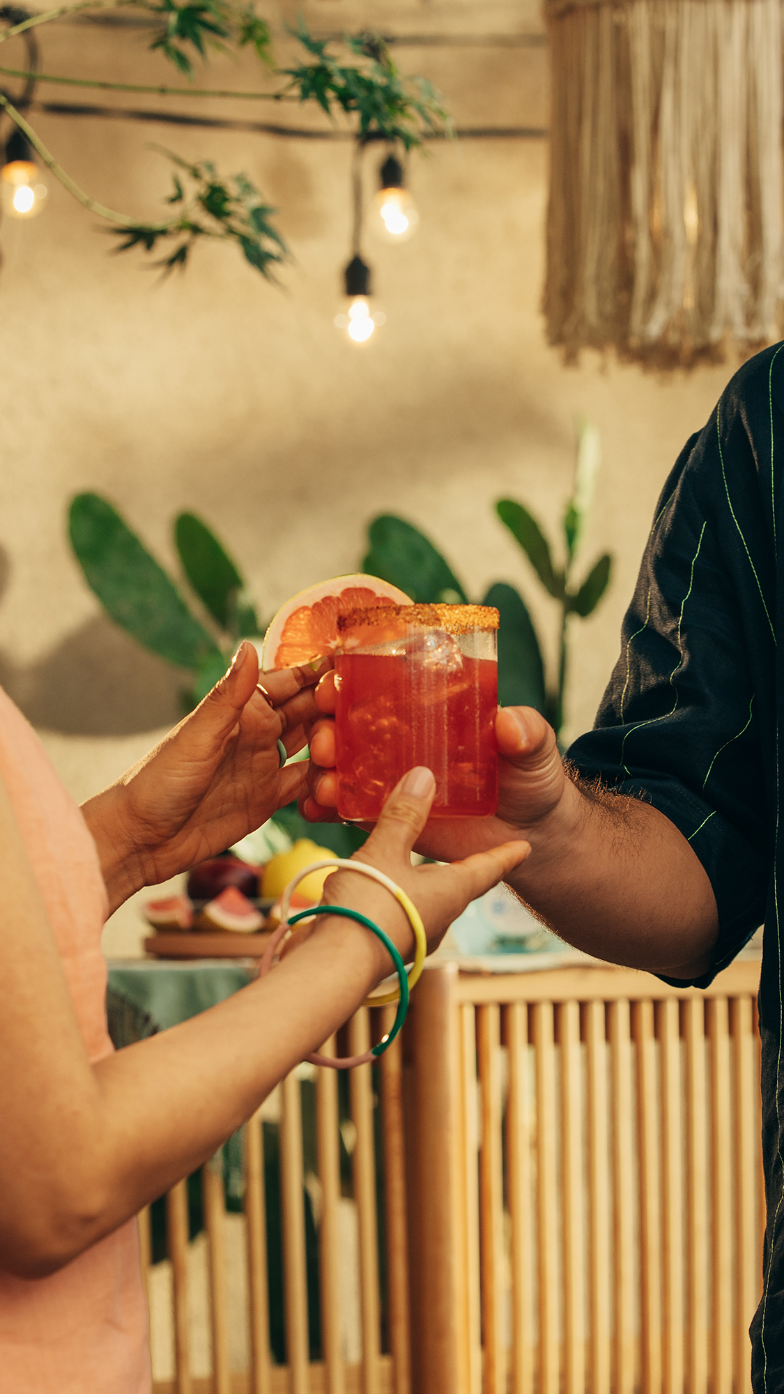 Una persona entrega a otra un vaso lleno del cóctel Blood Orange Paloma adornado con una naranja sanguina