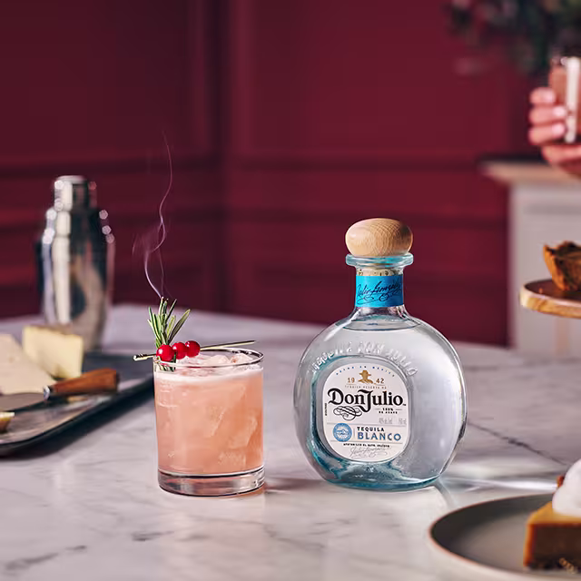 A bottle of Don Julio Blanco on a table next to a glass filled with Cranberry Rosemary Margarita cocktail