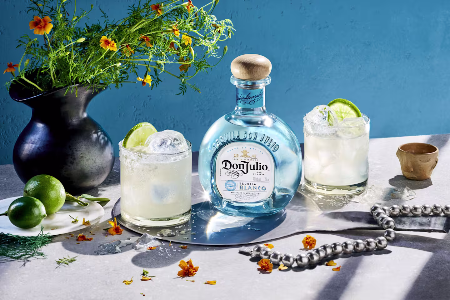 Image of a bottle of Don Julio Tequila Blanco displayed on a table with two cocktails in short glasses, each garnished with lime wedges.