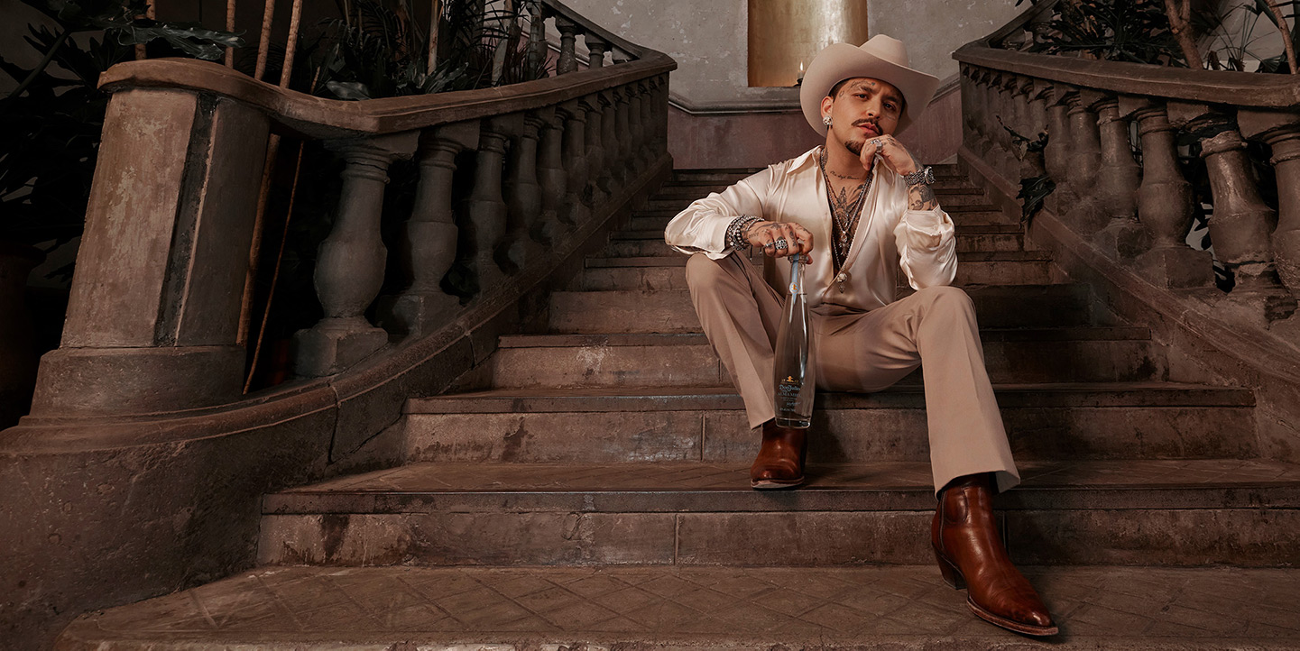 Mexican Singer-Songwriter Christian Nodal sitting on stairs holding a bottle of Don Julio Alma Miel Tequila.