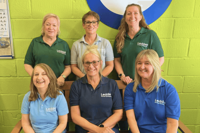 Picture showing the Loddon Social Enterprise Team. A group of six people stood in two rows in front of a bright green wall wearing Loddon Social Enterprise branded t-shirts and smiling.